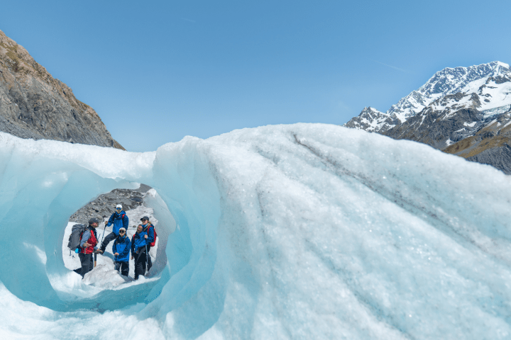Group of hikers inside an ice tunnel on a glacier with mountains in the background.
