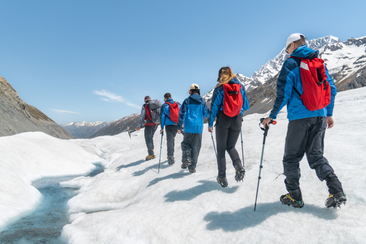 Group of hikers with red backpacks walking on a snowy mountain path, blue sky in background.