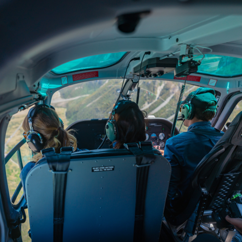 Inside view of a helicopter cockpit with three people wearing headsets, flying over a scenic landscape.