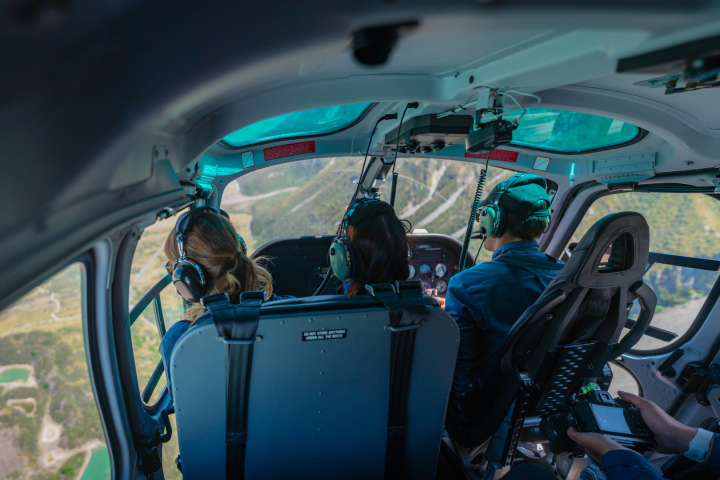 Inside view of a helicopter cockpit with three people wearing headsets, flying over a scenic landscape.