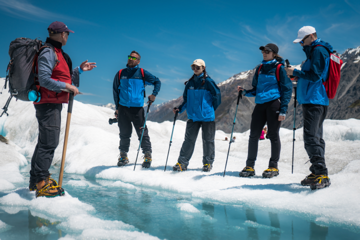 Group of hikers with guide on a glacier, wearing crampons, clear sky, and snowy mountains in background.