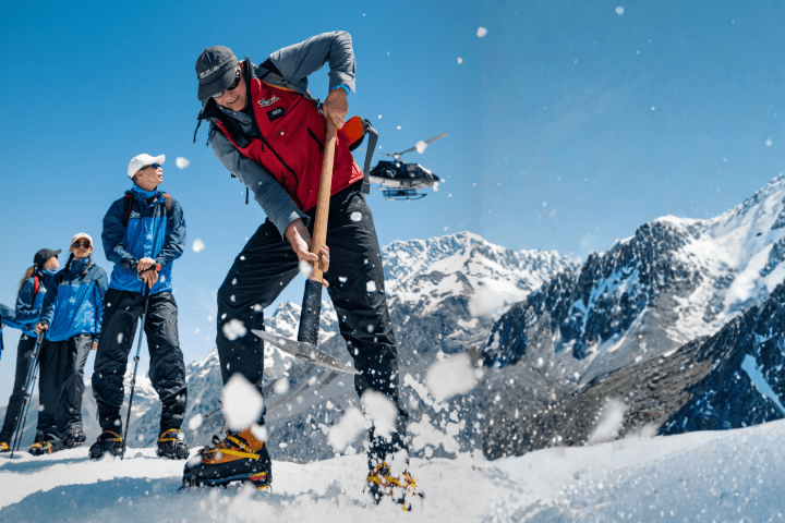 Group of climbers on a snowy mountain, one using an ice axe, with a helicopter nearby in the sky.