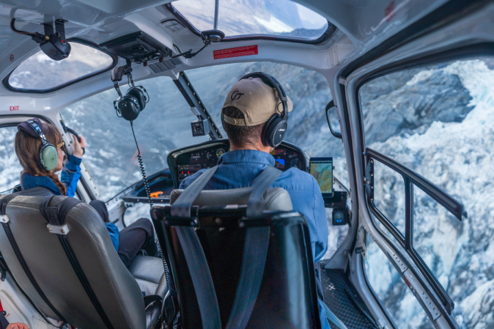 Interior of a helicopter with two pilots flying over a snowy mountain landscape.