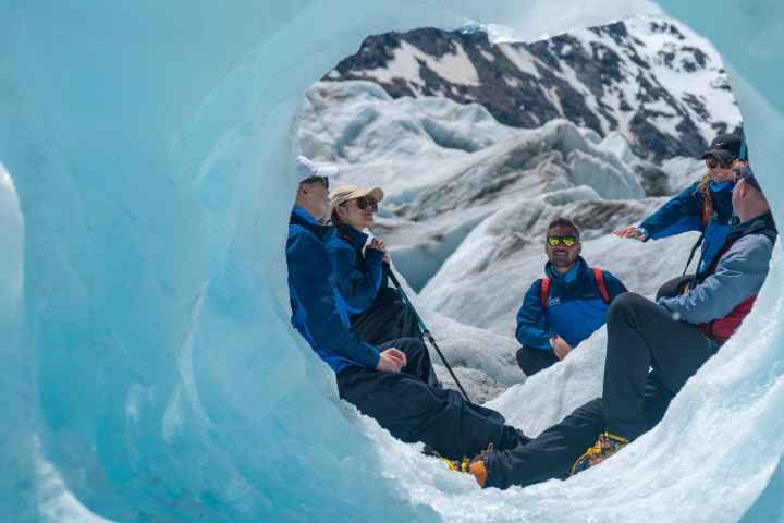 People sitting in an ice cave with snowy mountains in the background.