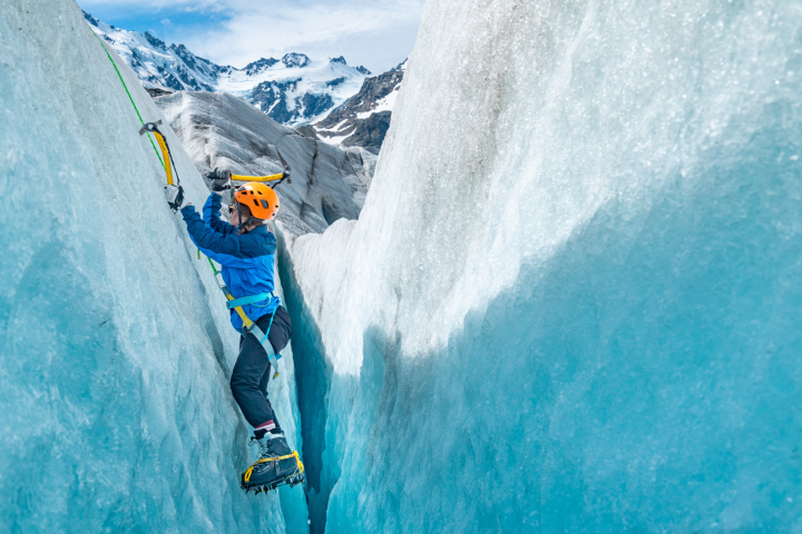 Ice climber in blue gear scaling a crevasse with snow-covered mountains in the background.