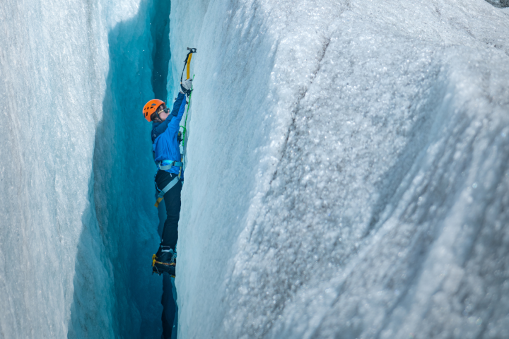 Climber ascends icy crevasse using ice axe, wearing a helmet and crampons.