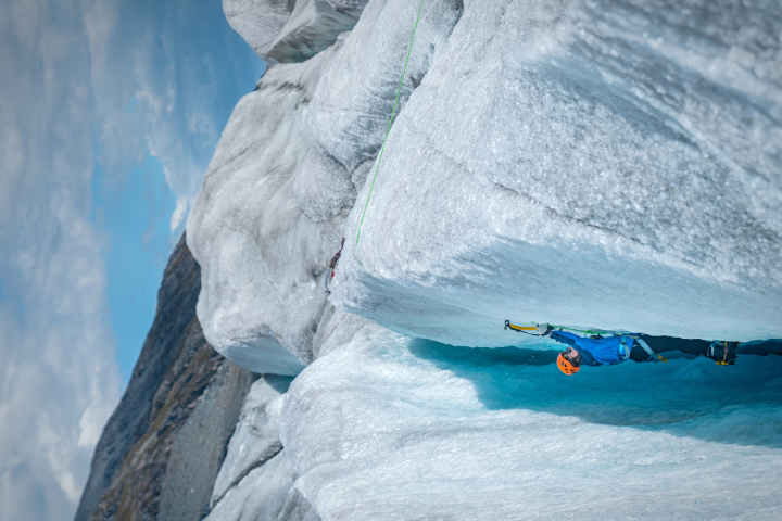 Climber wearing helmet ascends a steep ice wall using ice axes against a cloudy sky.