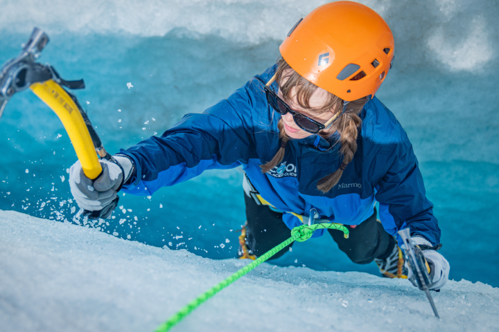 Person in orange helmet ice climbing with axe and rope on snowy surface.