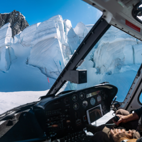 Helicopter cockpit view of pilot flying near snow-covered mountains and blue sky.