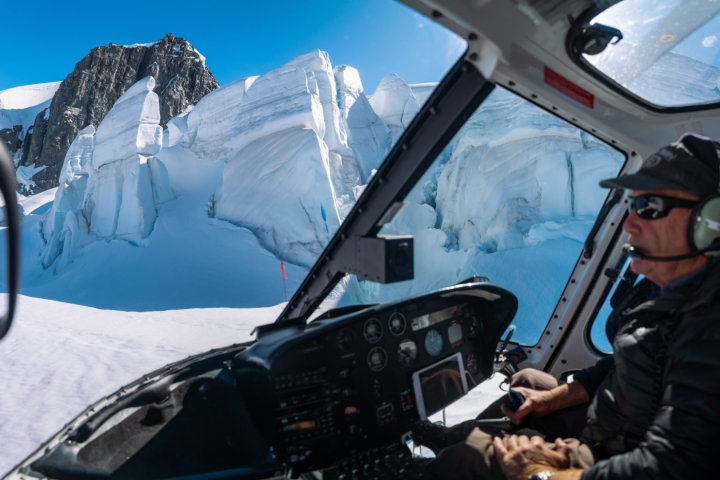 Helicopter cockpit view of pilot flying near snow-covered mountains and blue sky.