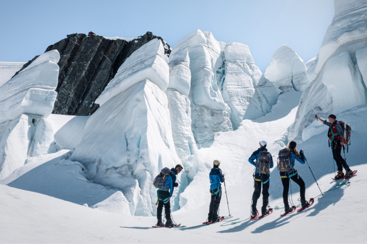 Group of five skiers with poles ascending snowy mountain with large ice formations.