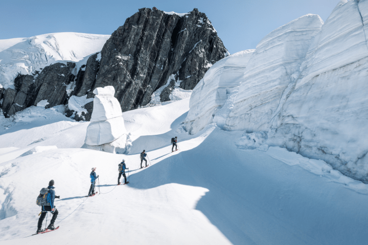 Five climbers traverse snow near large ice formations and rocky peaks.
