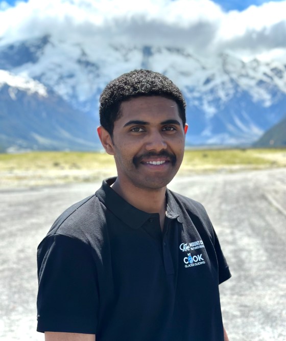 Person smiling in a black shirt with snowy mountains and blue sky in the background.