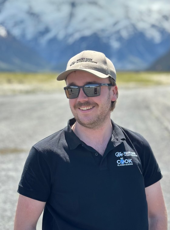 Smiling person in sunglasses and cap with mountains and snow in the background.