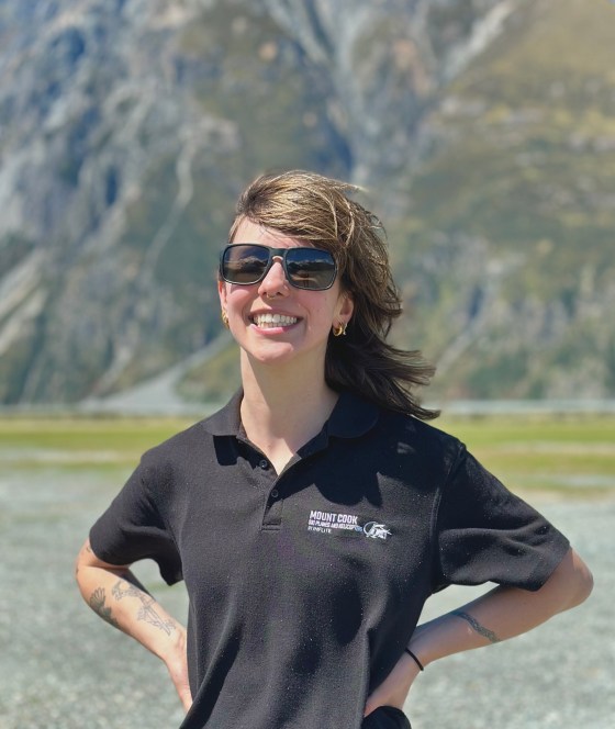 Person in sunglasses smiling outdoors with mountains in the background.