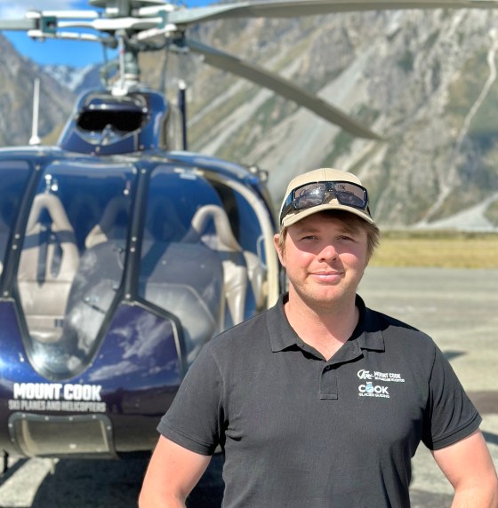 Person in a black shirt and cap stands in front of a helicopter with mountains in the background.