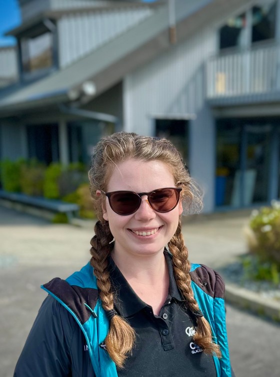 Smiling person with sunglasses and braids, standing outside a building on a sunny day.