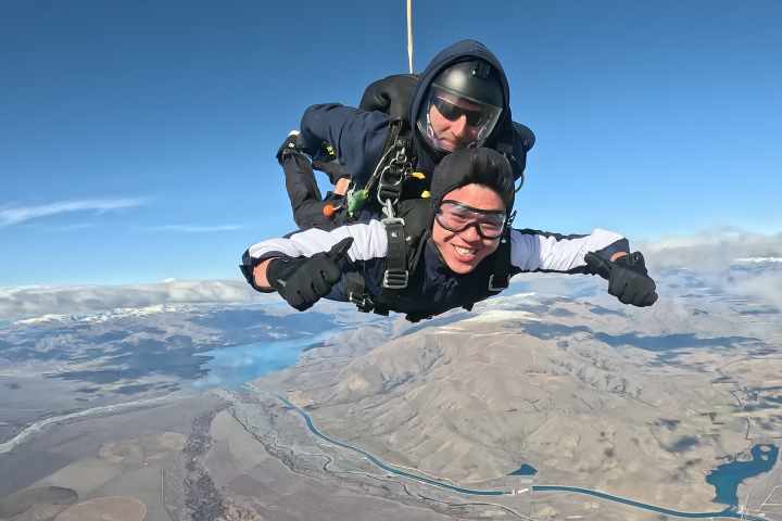 Two people tandem skydiving with a scenic mountain and river landscape below.