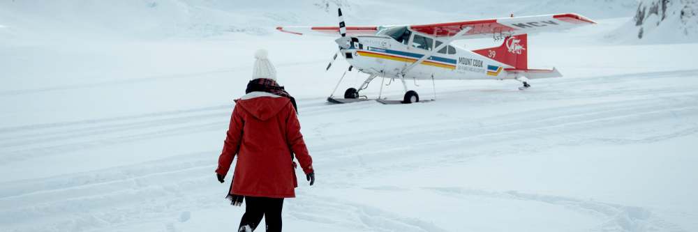 Person in red coat approaches small plane on snowy terrain.