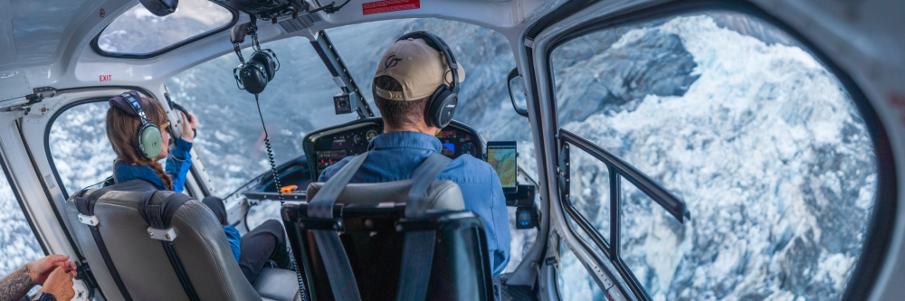 Two pilots in a helicopter cockpit flying over snowy mountains.