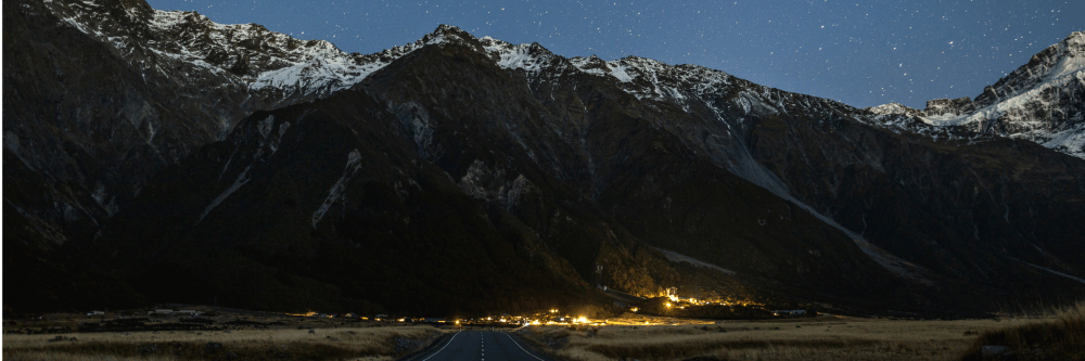 Mountainous landscape at night with starry sky and distant village lights.
