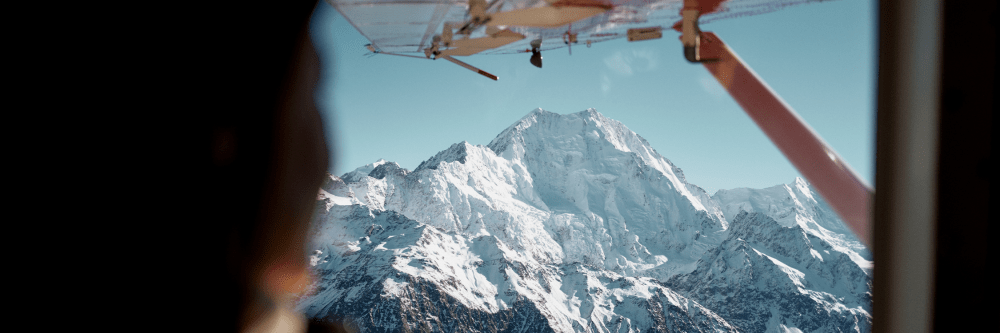 View of snowy mountain from airplane window with wing visible.