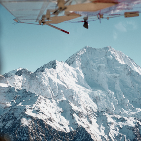 View of snowy mountain from airplane window with wing visible.