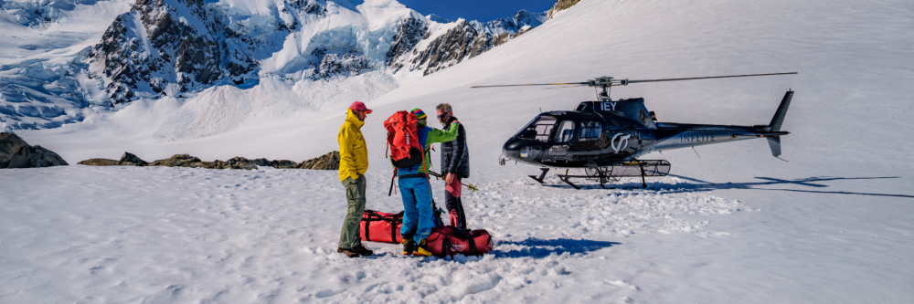 Three people on snow with gear near a helicopter in a mountainous area.