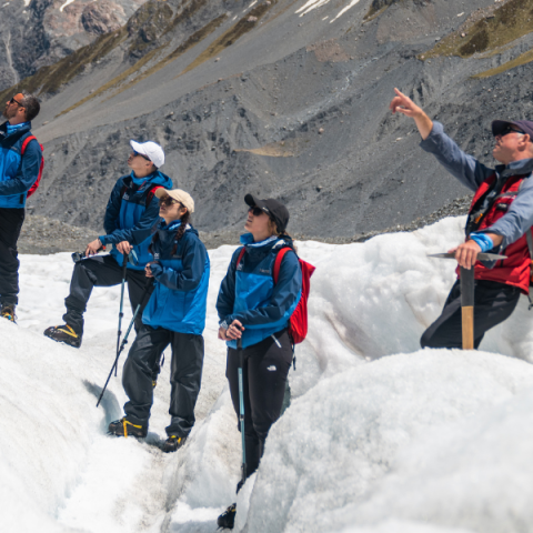 Four hikers on snowy terrain with mountains in the background.