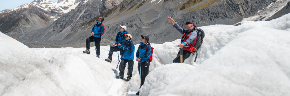 Four hikers on snowy terrain with mountains in the background.