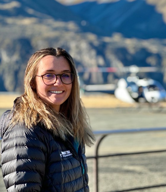 Woman in glasses smiling with a helicopter and mountains in the background.