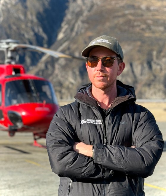 Man in sunglasses and cap stands confidently in front of a red helicopter.